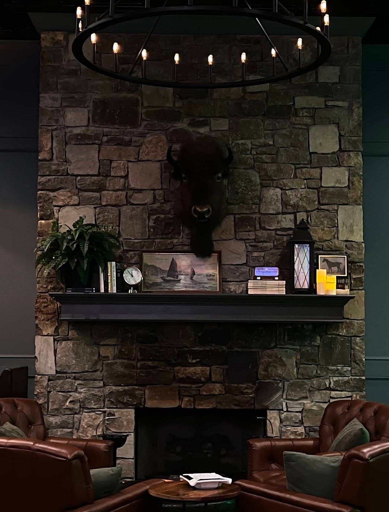 Stone fireplace at The Royale with a bison mount above the mantel, an iron chandelier, and leather chairs